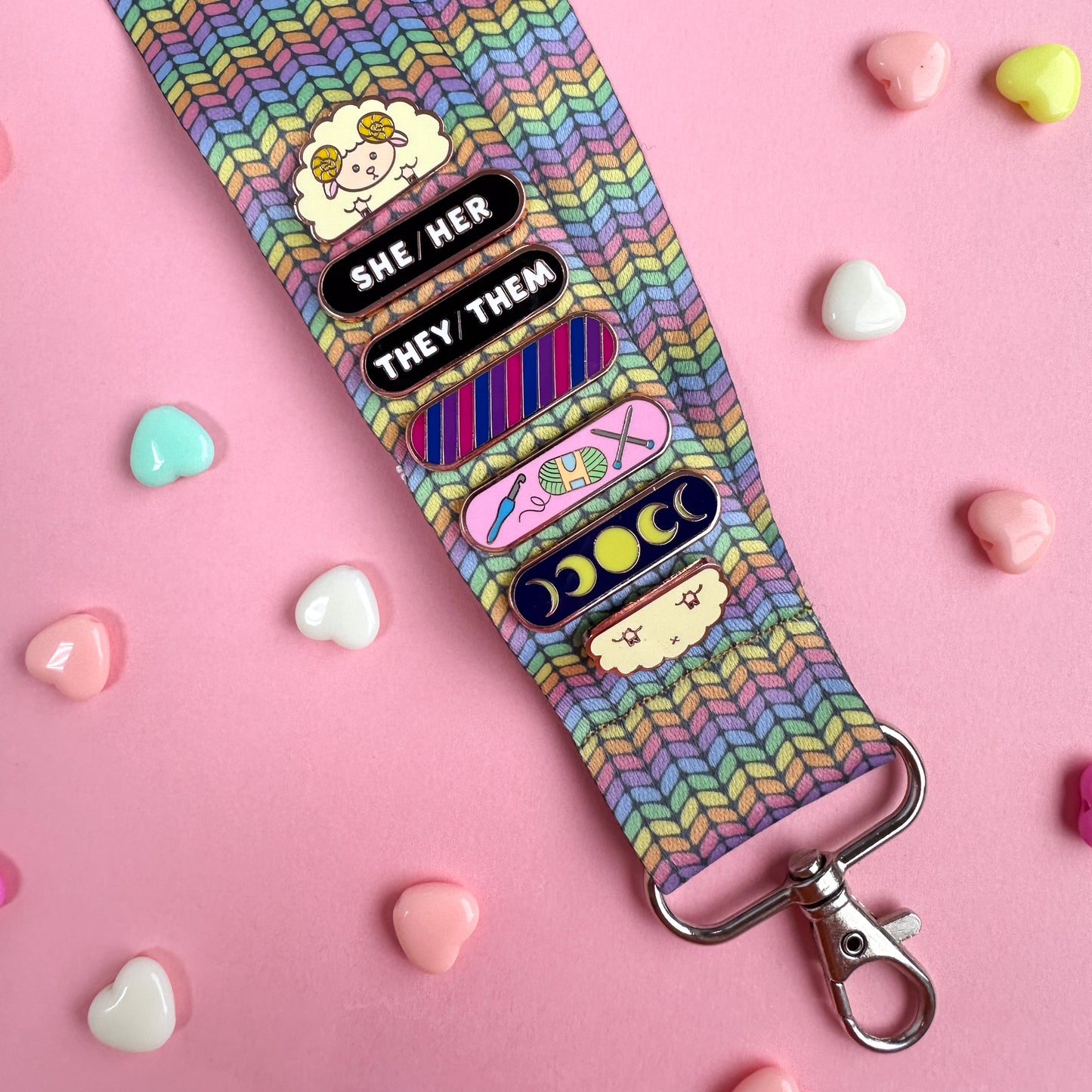A woven lanyard with a pastel rainbow stockinette stitch graphic printed on it with a silver metal lobster claw clasp at the bottom. The lanyard has a Sheep Pride Pal pin displayed on it with She/Her and They/Them pronoun plaques stacked with a Bi Pride Plaque, Crafts Personality Plaque, and Moon Phases Personality Plaque. 