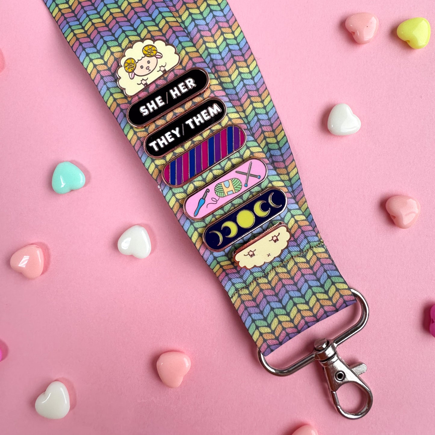 A woven lanyard with a pastel rainbow stockinette stitch graphic printed on it with a silver metal lobster claw clasp at the bottom. The lanyard has a Sheep Pride Pal pin displayed on it with She/Her and They/Them pronoun plaques stacked with a Bi Pride Plaque, Crafts Personality Plaque, and Moon Phases Personality Plaque.