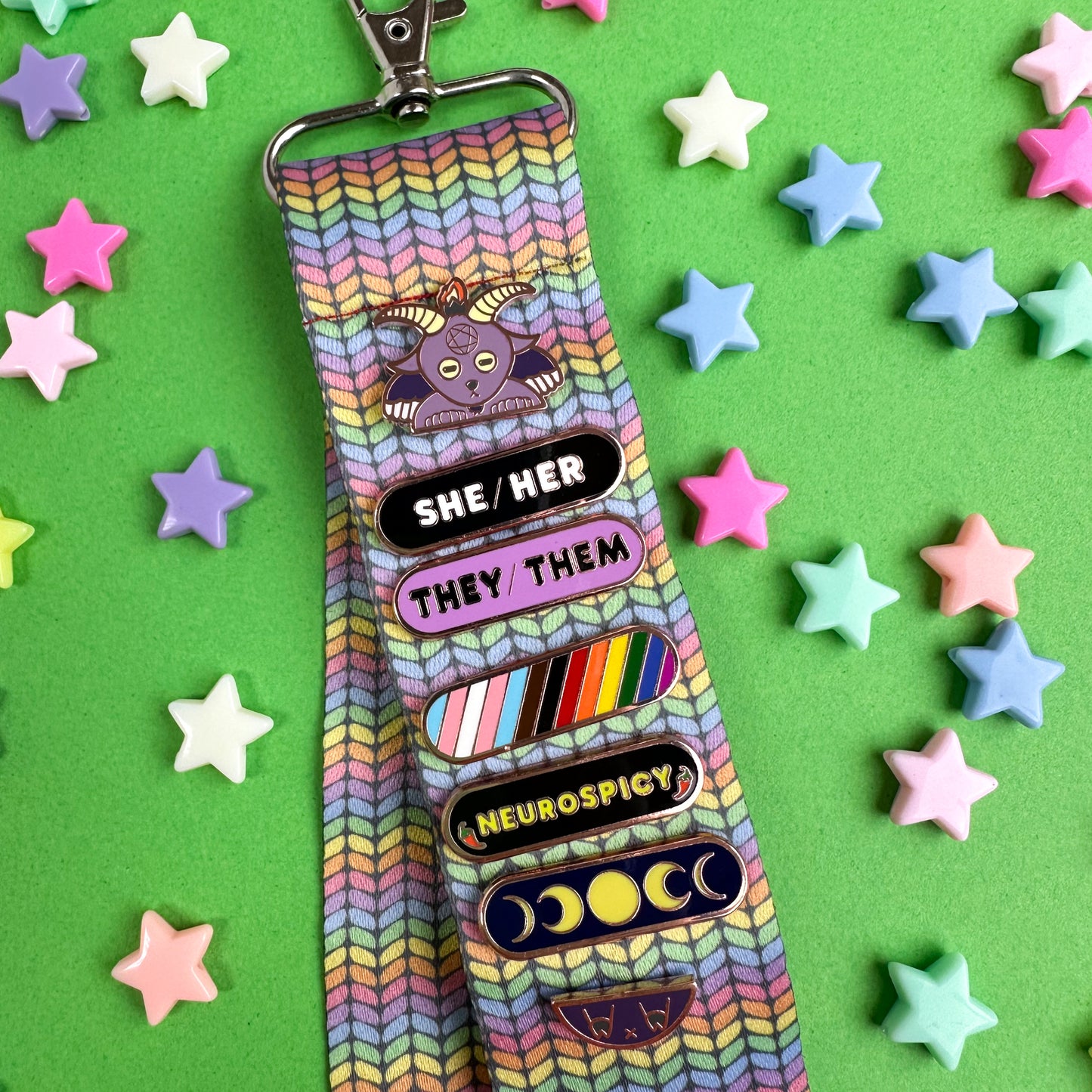 A woven lanyard with a pastel rainbow stockinette stitch graphic printed on it with a silver metal lobster claw clasp at the bottom. The Lanyard has a Baphomet Pride Pal displayed on it with a black She/Her Plaque, a purple They/Them plaque, an inclusive Pride Plaque, a Neurospicy Plaque, and a Moon Phases Plaque.