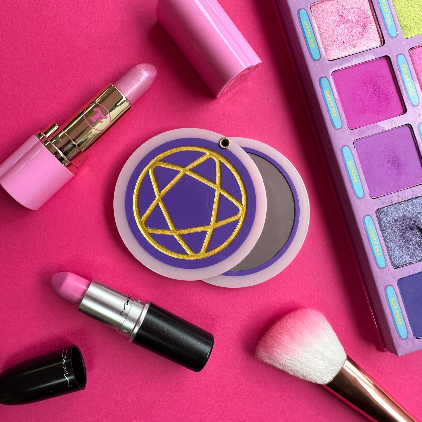 A circular hanged compact mirror that is a purple circle with a pastel pink border with a gold shiny pentacle embossed on the cover. It is on a pink background with two open pink lipsticks, a pink makeup brush and bright purple eyeshadow palette surrounding it.