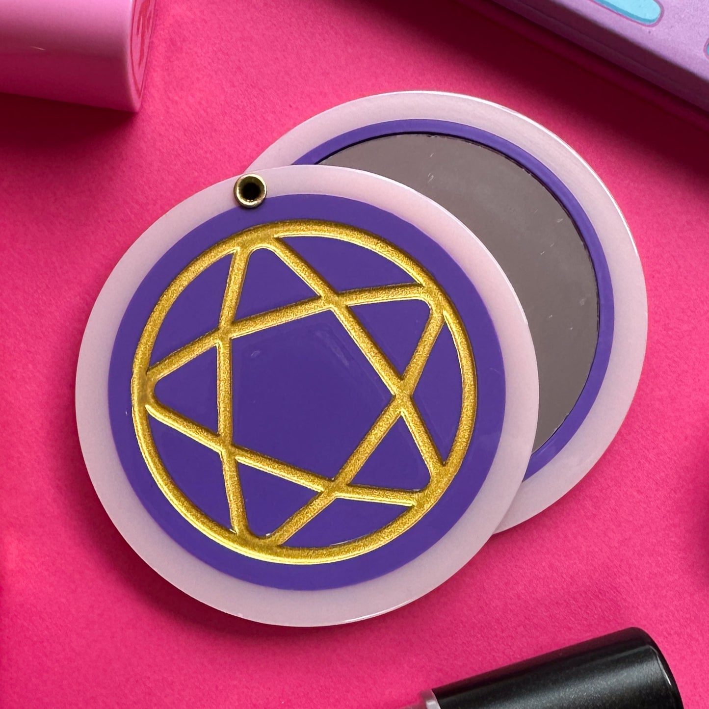 A circular hanged compact mirror that is a purple circle with a pastel pink border with a gold shiny pentacle embossed on the cover. The mirror is on a hot pink background with some makeup around it.