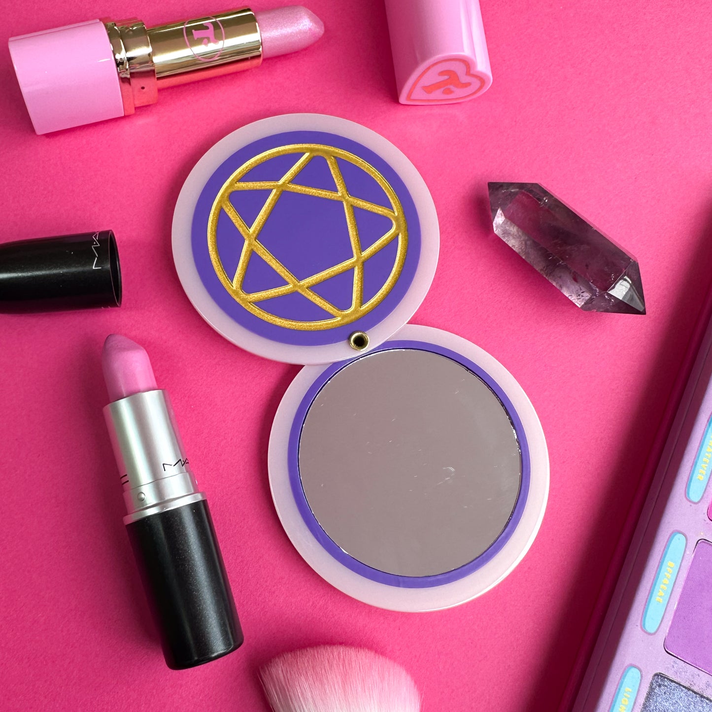 The pentacle mirror all the way open to show the full circular mirror inside the two pink and purple circles. There is a crystal and open lipsticks around the mirror.