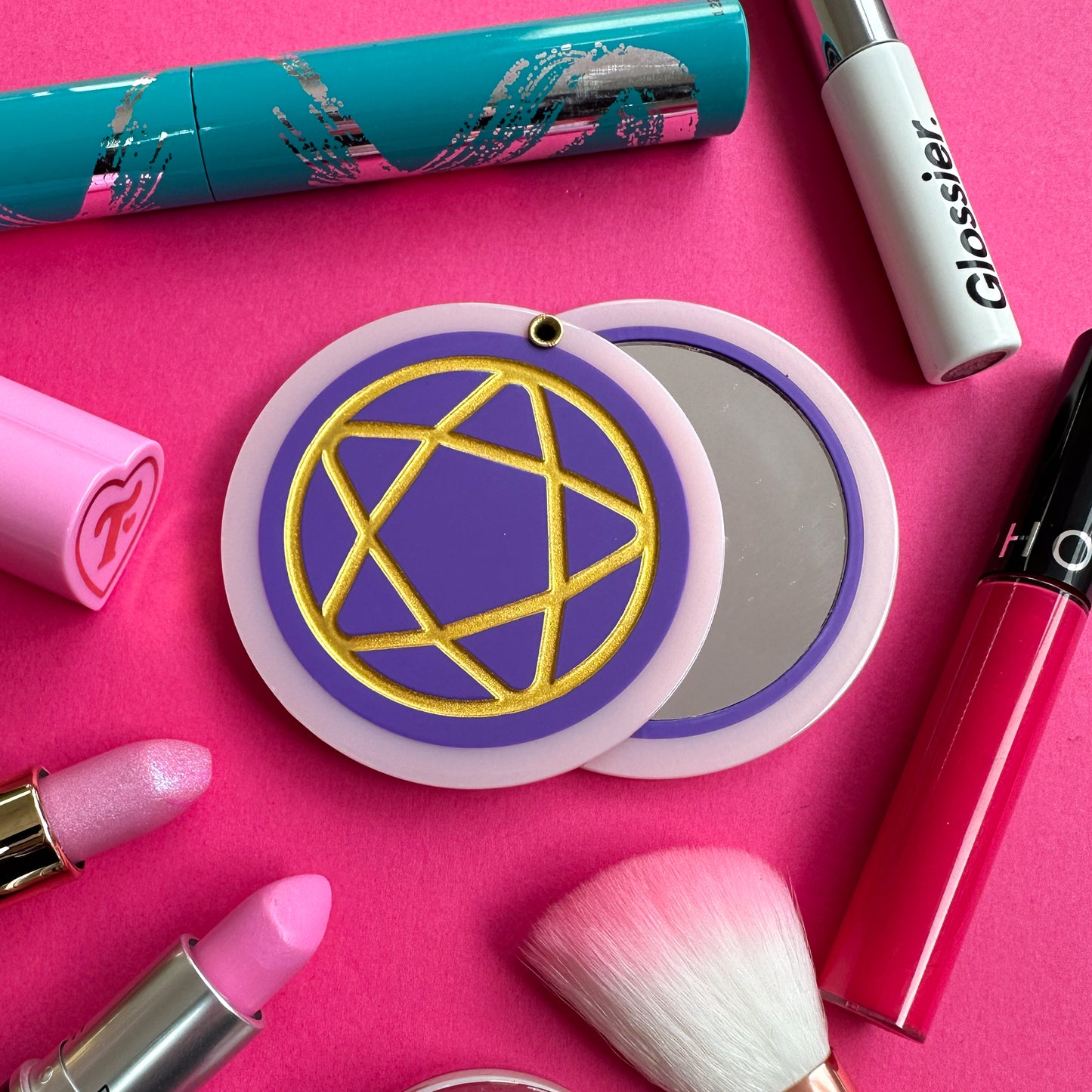 A circular hanged compact mirror that is a purple circle with a pastel pink border with a gold shiny pentacle embossed on the cover. There are various lipsticks and mascara tubes around the mirror.