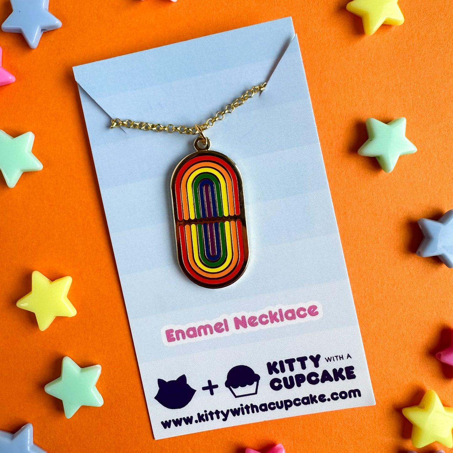 A gold pendant style necklace with an oval pendant with mirrored rainbows on it. The necklace is packaged on a blue card. The card is on an orange background with plastic stars around it.