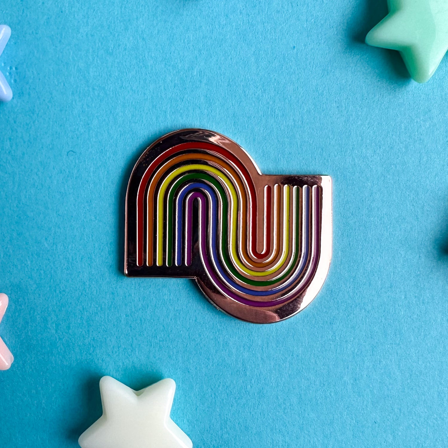 An enamel pin with one right side up rainbow intersecting with an upside down rainbow on copper metal. The pin is on a blue piece of paper with plastic star beads around it.