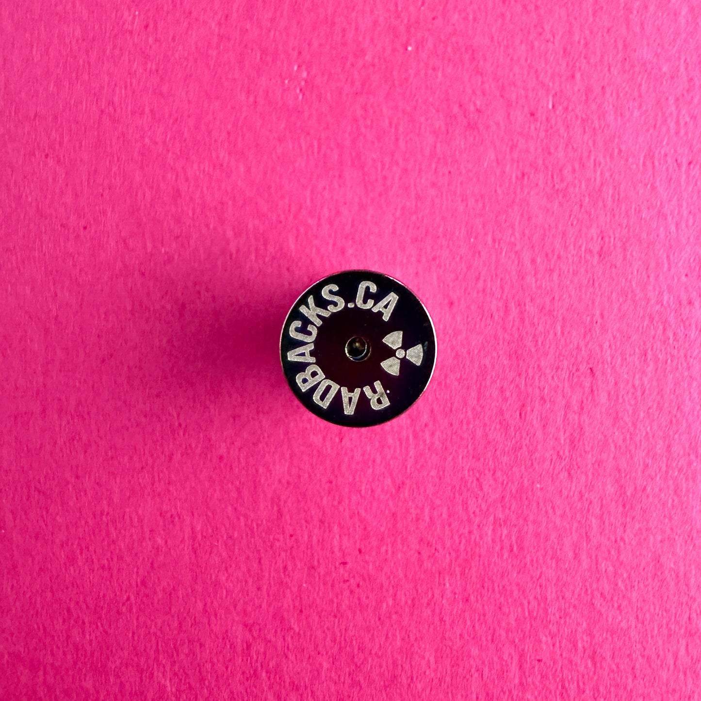 A metal pin back with etched circular text reading "Radbacks.ca" with an atomic symbol. The pin back is on a pink paper background.