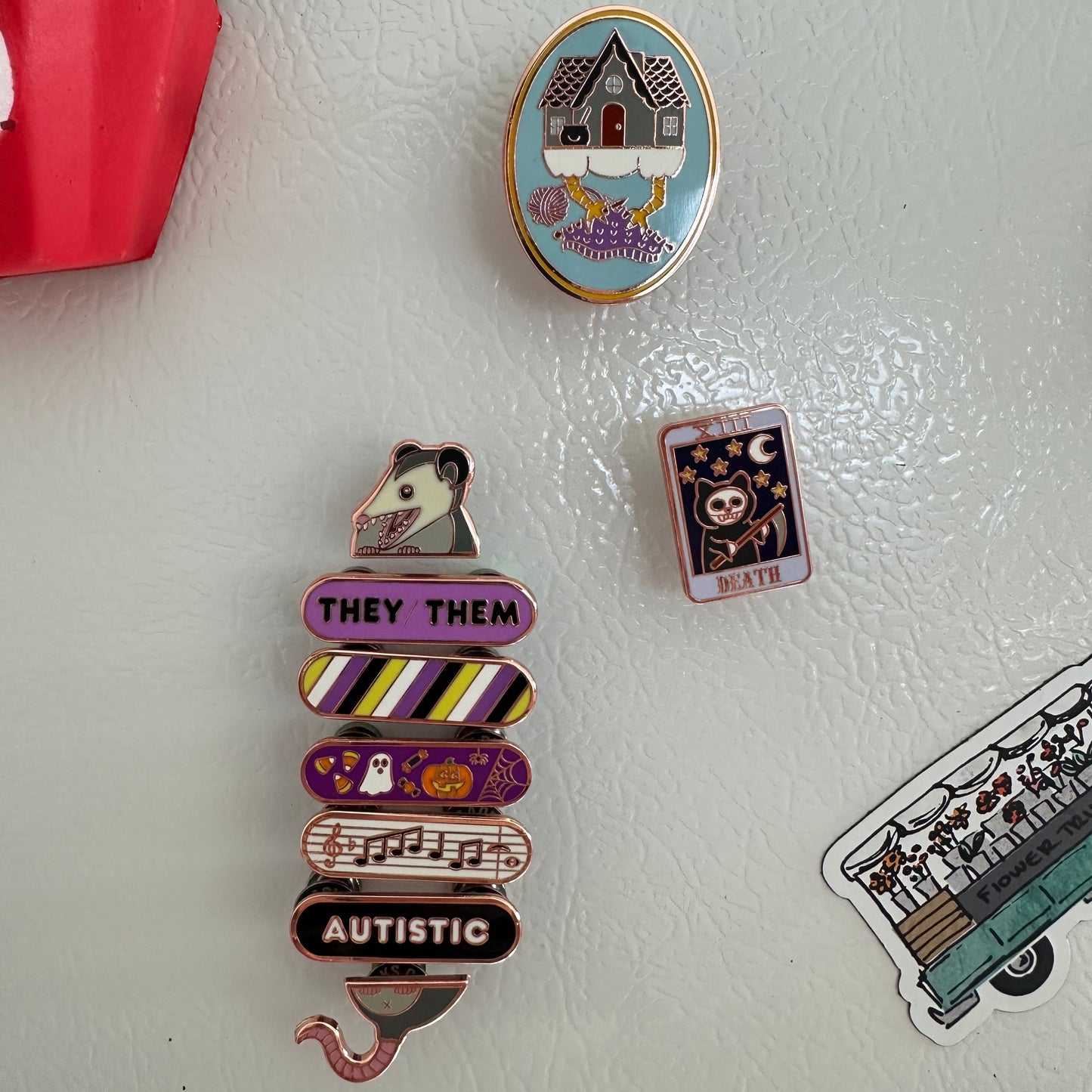 The side of a fridge with enamel pins stuck to it like magnets. There is a baba yaga pin, a death tarot card kitty pin, and a Pride Pal Stack with an opossum and the nonbinary pride flag.