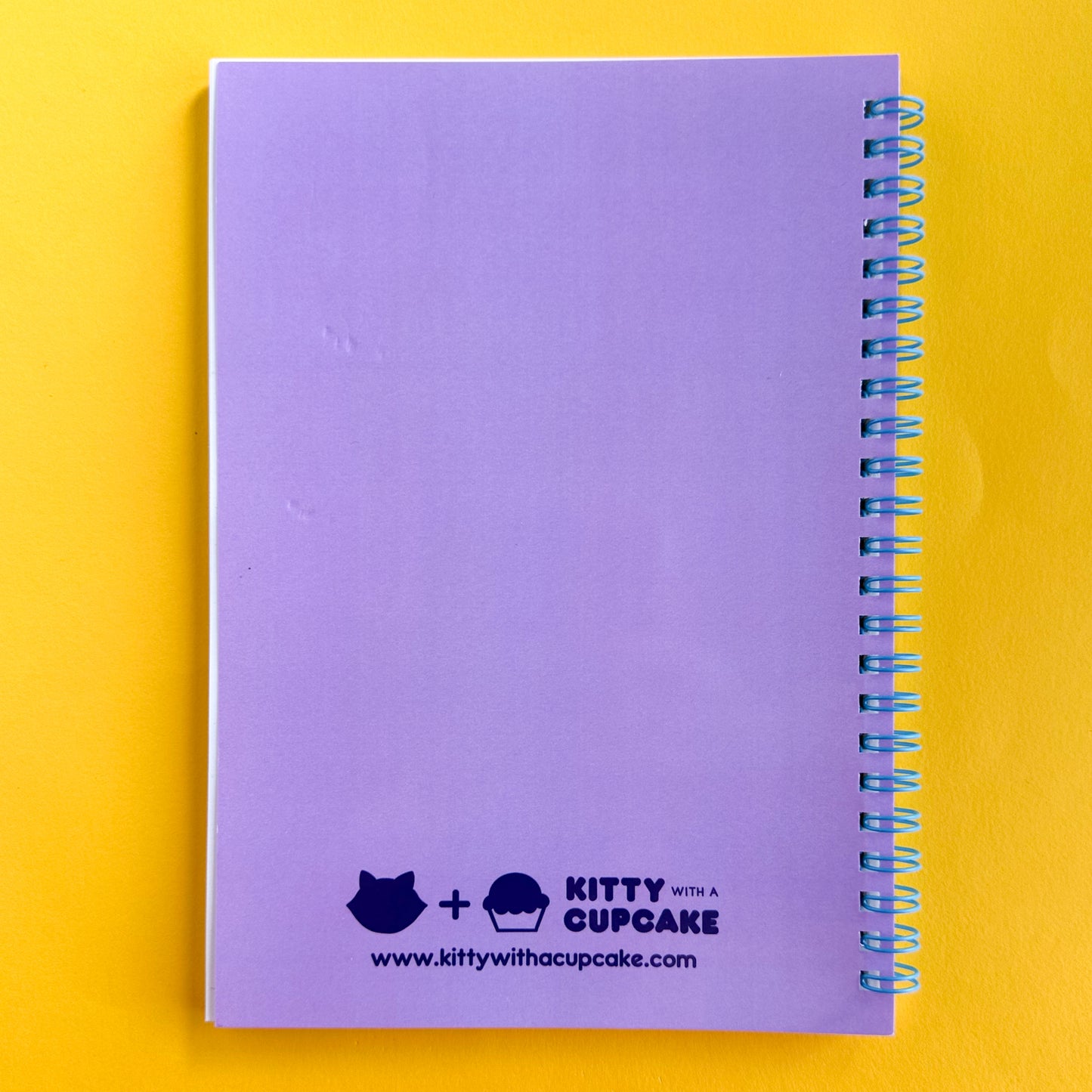 A spiral notebook with a lavender back cover with a Kitty With A Cupcake Logo and www.kittywithacupcake.com printed at the bottom center in dark purple bubble font. The notebook is sitting on a yellow paper background.