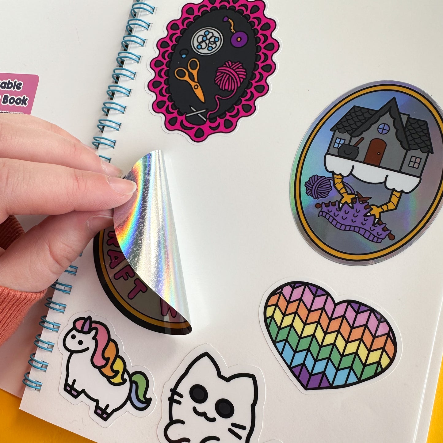 A hand peeling up a holographic stick from a page of a reusable sticker book. There are several cute illustrated stickers in the book including a sticker of a pastel rainbow heart with a knit pattern, chubby rainbow unicorn, and baba Yaga's house knitting.