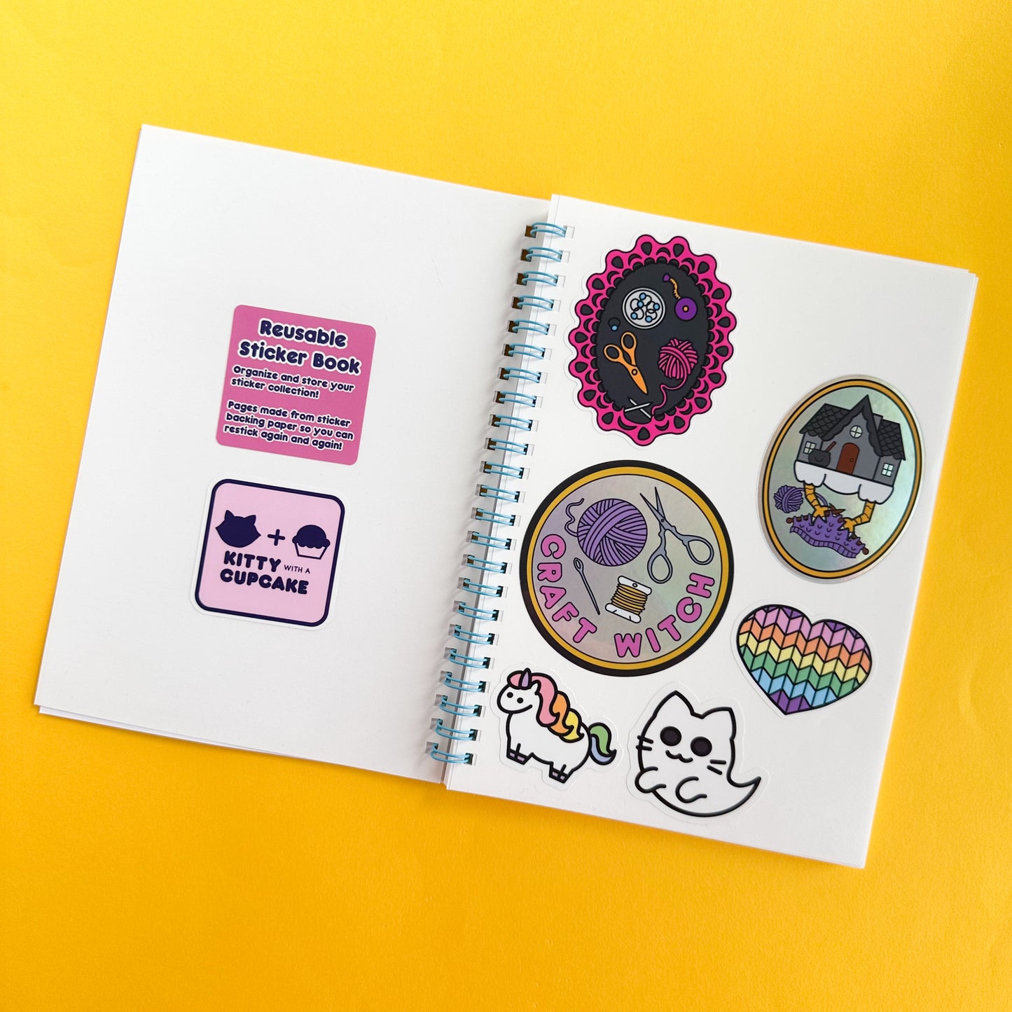 A spiral notebook open with stickers on both pages. The stickers are in a cute cartoony illustrative style and have cute characters like a chubby unicorn and ghost kitty. One of the stickers reads "Reusable Sticker Book. Oraganize and store your sticker collection! Pages made from sticker backing paper so you can restick again and again!" The spiral of the notebook is light blue and the notebook is sitting on a yellow paper backdrop.