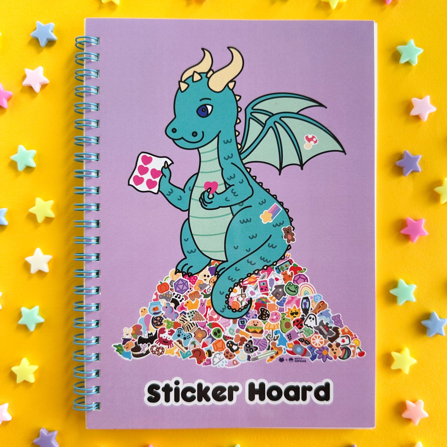 A spiral bound notebook with a lavender cover with an illustration of a teal dragon holding stickers and sitting on top of a giant pile of stickers. Underneath the dragon are the words "Sticker hoard". The notebook is on a yellow paper background with pastel plastic star pony beads strewn around it.