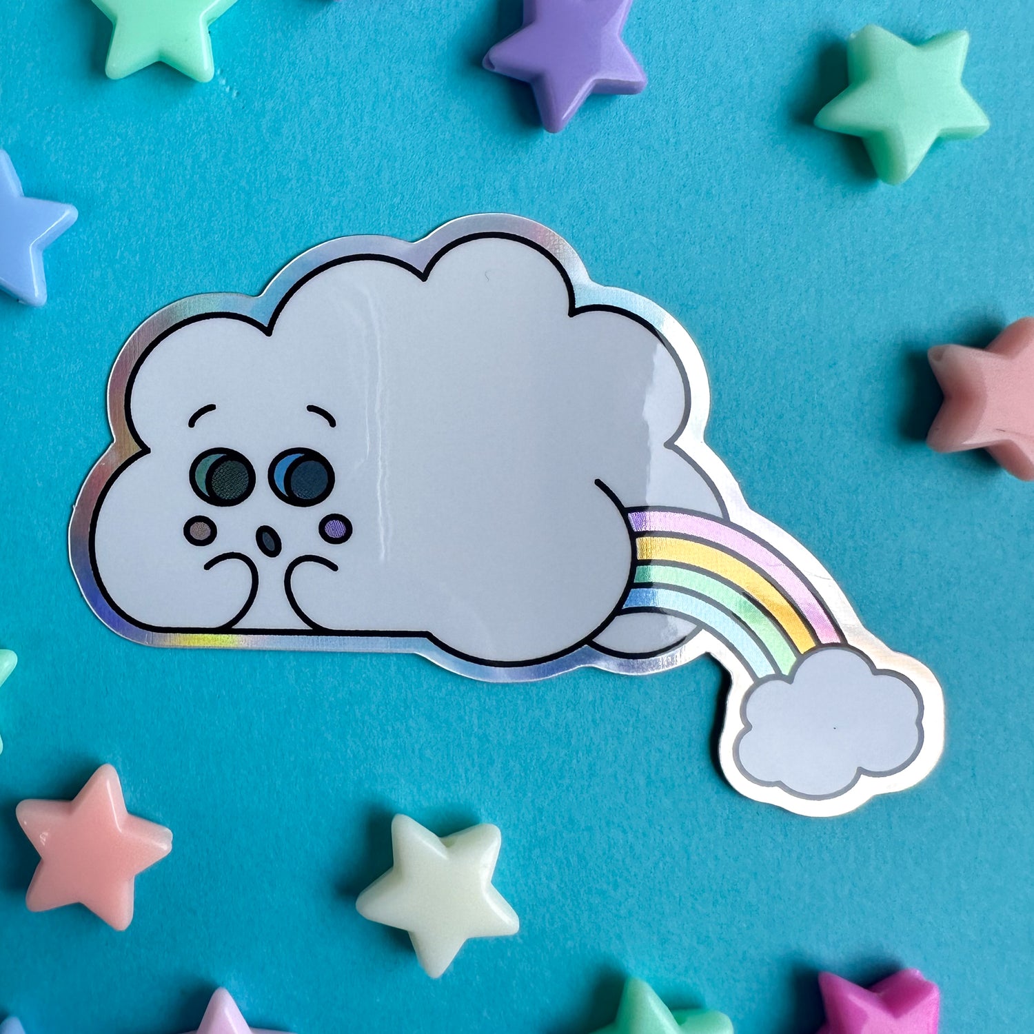 A holographic sticker shaped like a cloud with a cute face making an embarrassed expression, the cloud's lumps form a little booty that is farting out another cloud and rainbow.