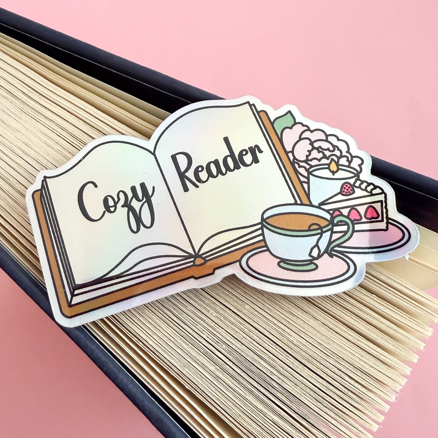A holographic sticker depicting an open book with “Cozy Reader” written across the pages. Beside the book are a full tea cup on a pink saucer, a slice of strawberry cake, a lit candle, and a baby pink peony with a green leaf. The sticker is displayed and the splayed edge of a book.