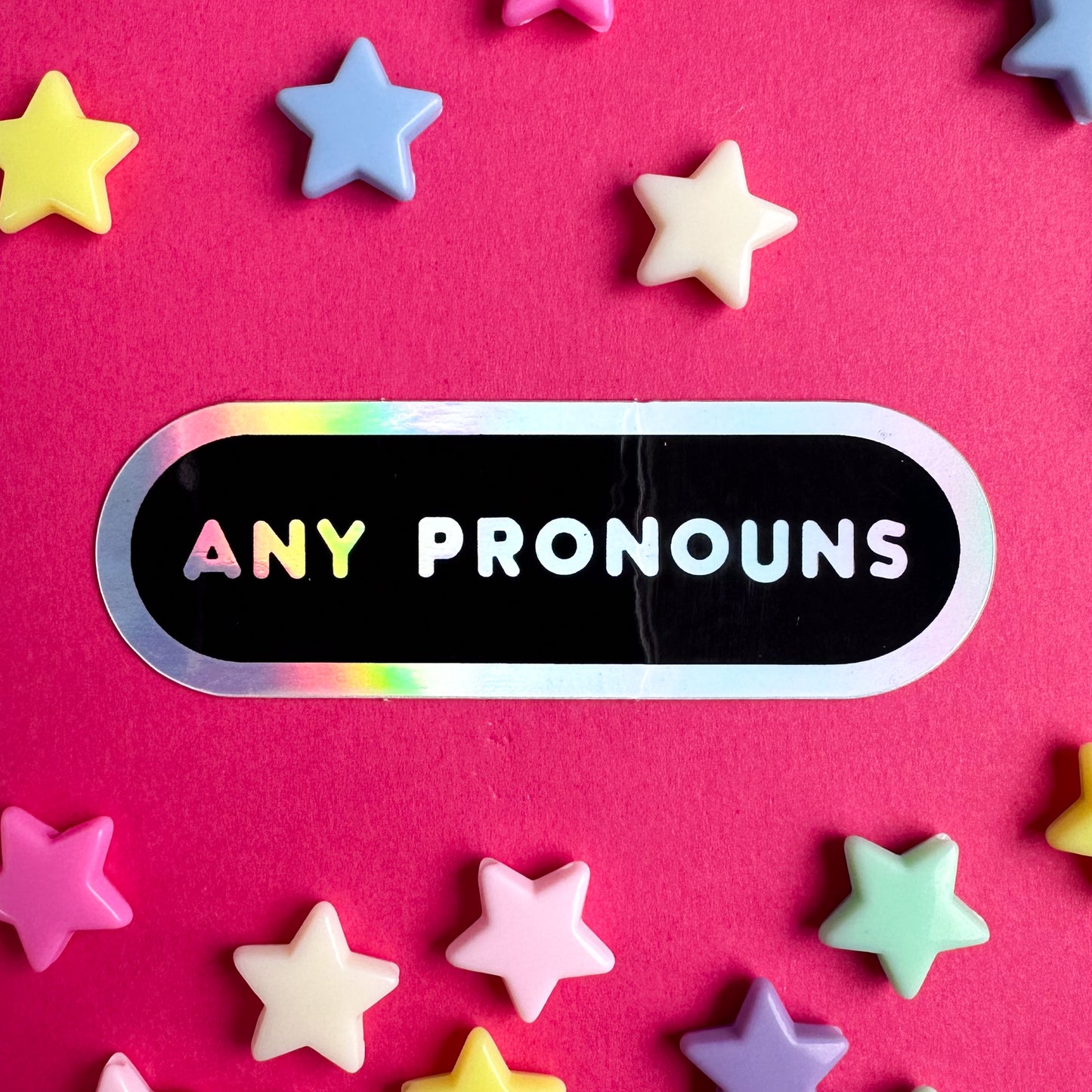 An oval sticker with the words "Any Pronouns" in holographic text. The sticker is sitting on a hot pink background with pastel stars around it.