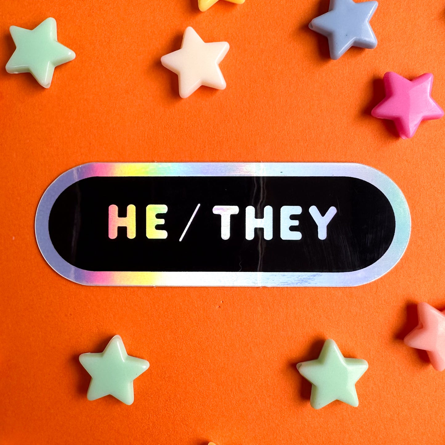 A capsule shaped sticker with holographic text that reads "He/They" on a black background. The sticker is on top of orange paper with star beads around it.