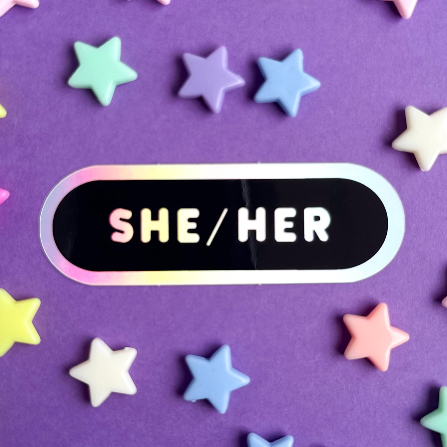 A sticker shaped like an oval that reads "She/Her" and is sitting on a purple background with star beads around it.