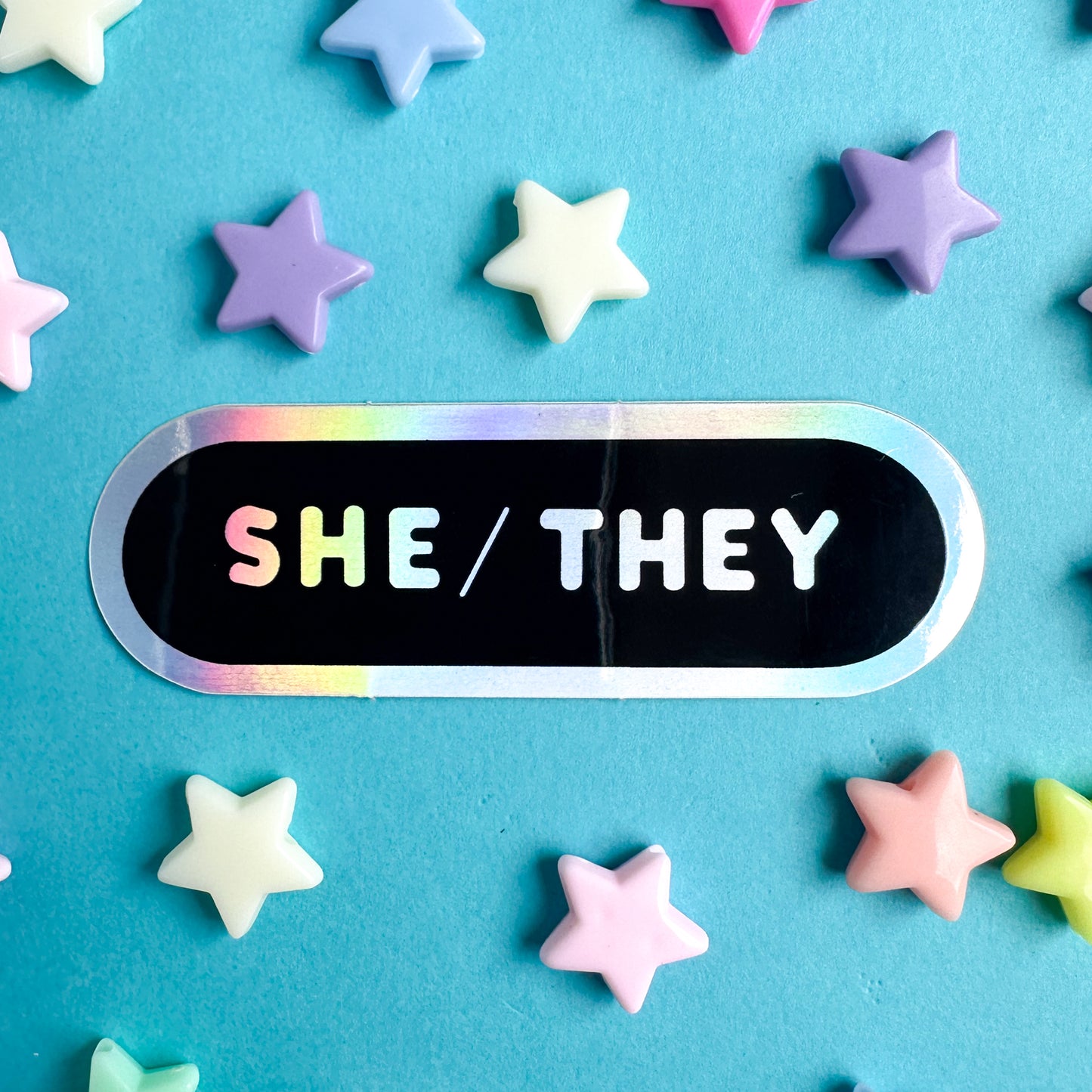 An oval shaped sticker with the words "She/They" in holographic letters on a blackground. The sticker is on a blue piece of paper with star beads around it.