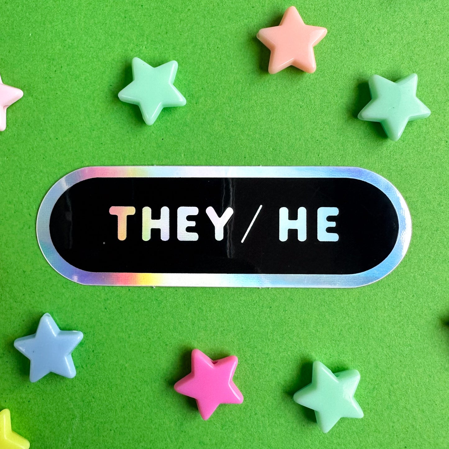 A black capsule shaped sticker with a holographic border and the pronouns They/He in holographic bubble letters. The sticker is on a green background with plastic star beads scattered around it.