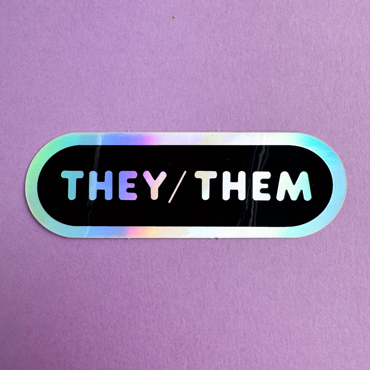 An oval shaped sticker with the words "They/Them" in holographic bubble letters on a black background. The sticker is on a purple piece of paper.