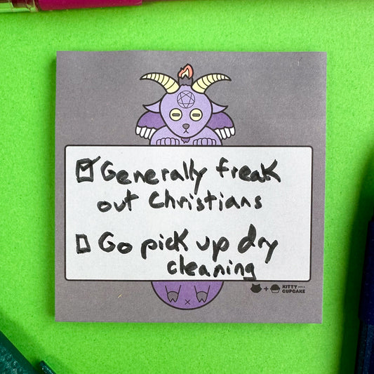 A charcoal grey square pad of sticky notes with an illustration of Baphomet on it holding a light grey box with a hand written to-do list in it. The list reads "Generally freak out Christians (checked), Go pick up dry cleaning (unchecked)"
