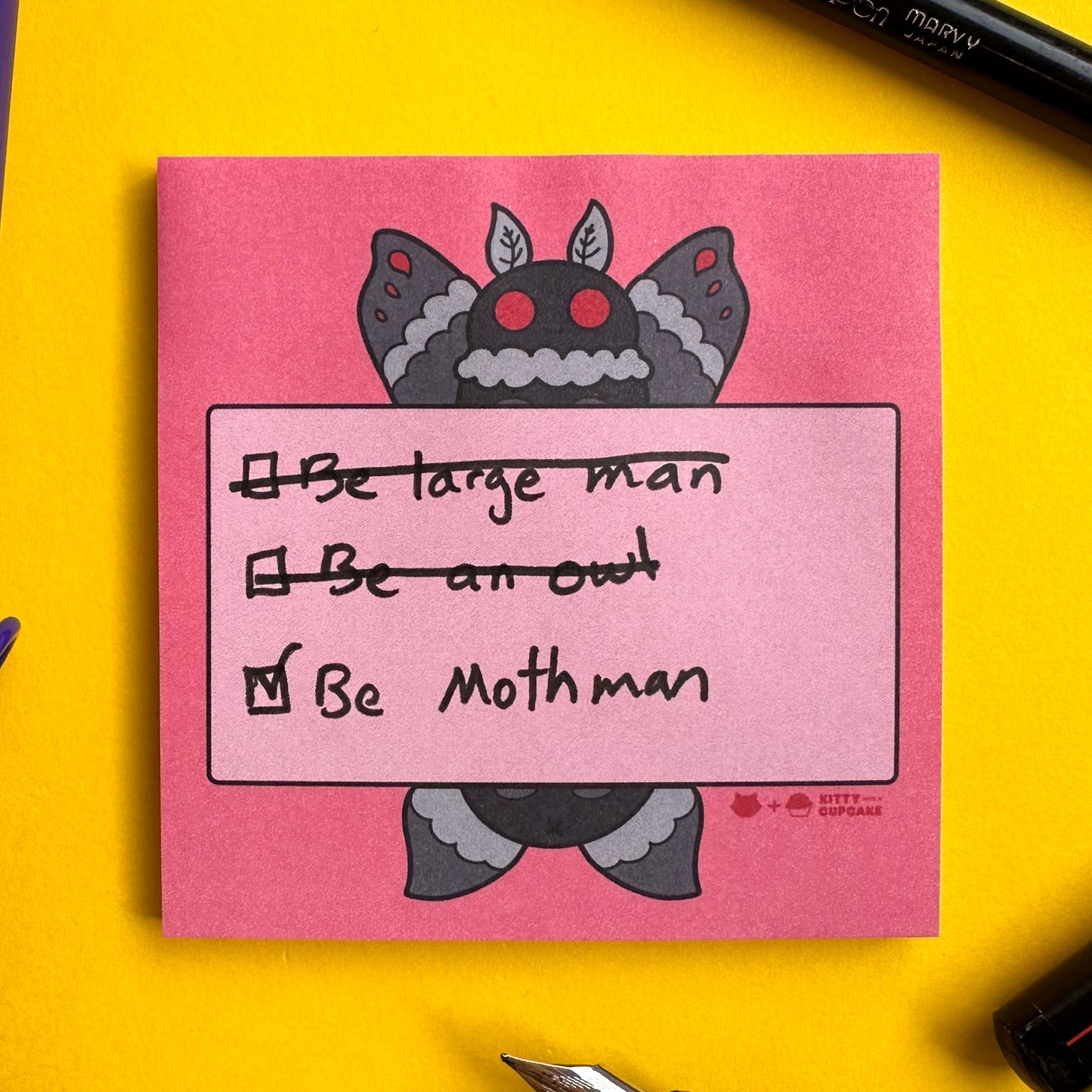A red square sticky note pad with an illustration of Mothman holding a pink box with a list written on it. The list reads "Be large man, Be an owl" which are both crossed out, and "Be Mothman" which is checked off. The notepad is on a yellow background with pens around it.