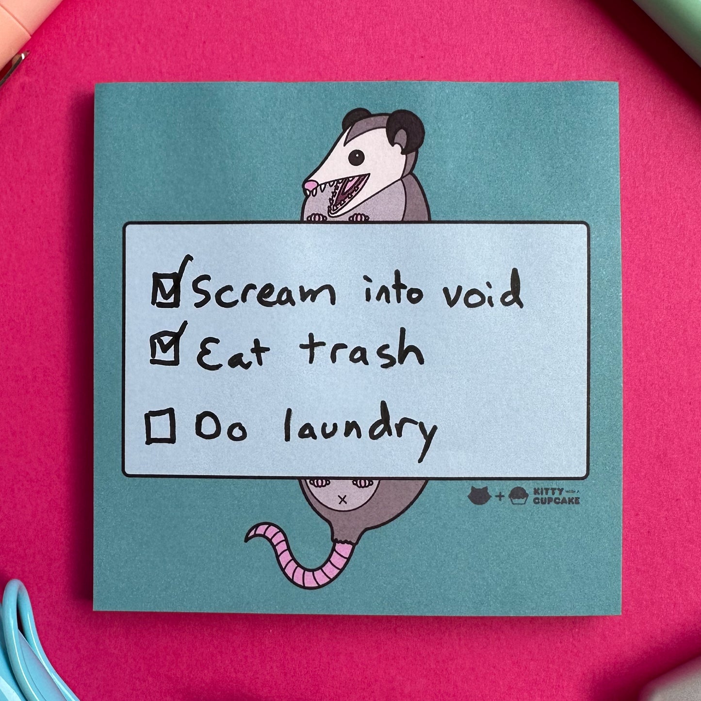 A blue square sticky note pad featuring an illustration of an opossum holding a light blue rectangle with a list written in it. The checklist reads "Scream into void, Eat Trash, Do Laundry" the first two items are checked off but the last is not. The sticky notes are on a hot pink background with some pens around them.