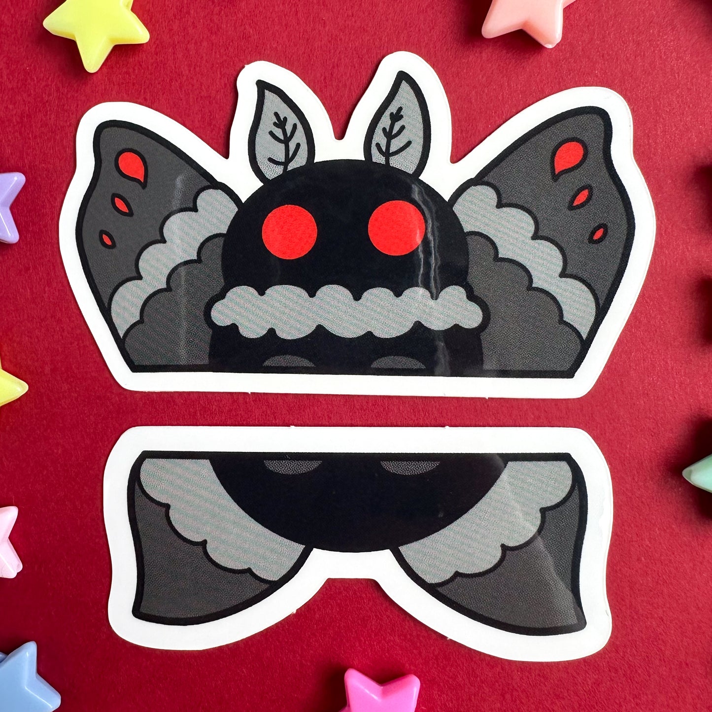 A pair of stickers that come together to form the top and bottom half of Mothman illustrated in a cartoony style with grey wings, a black body, and big red circle eyes. The stickers are on top of a red background with pastel star beads scattered around them.