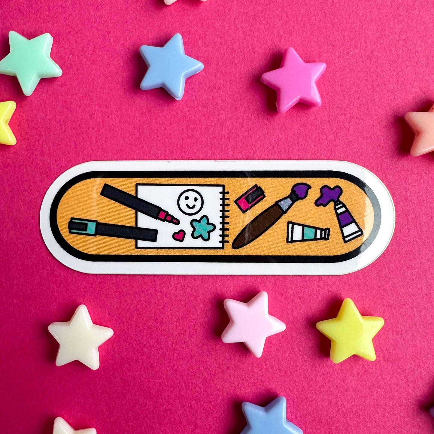 A vinyl sticker in the shape of an oval with illustrations of art supplies like paint pens, paintbrush, paints, and sketchbook on it. The sticker is on a pink paper background with pastel star beads around it.