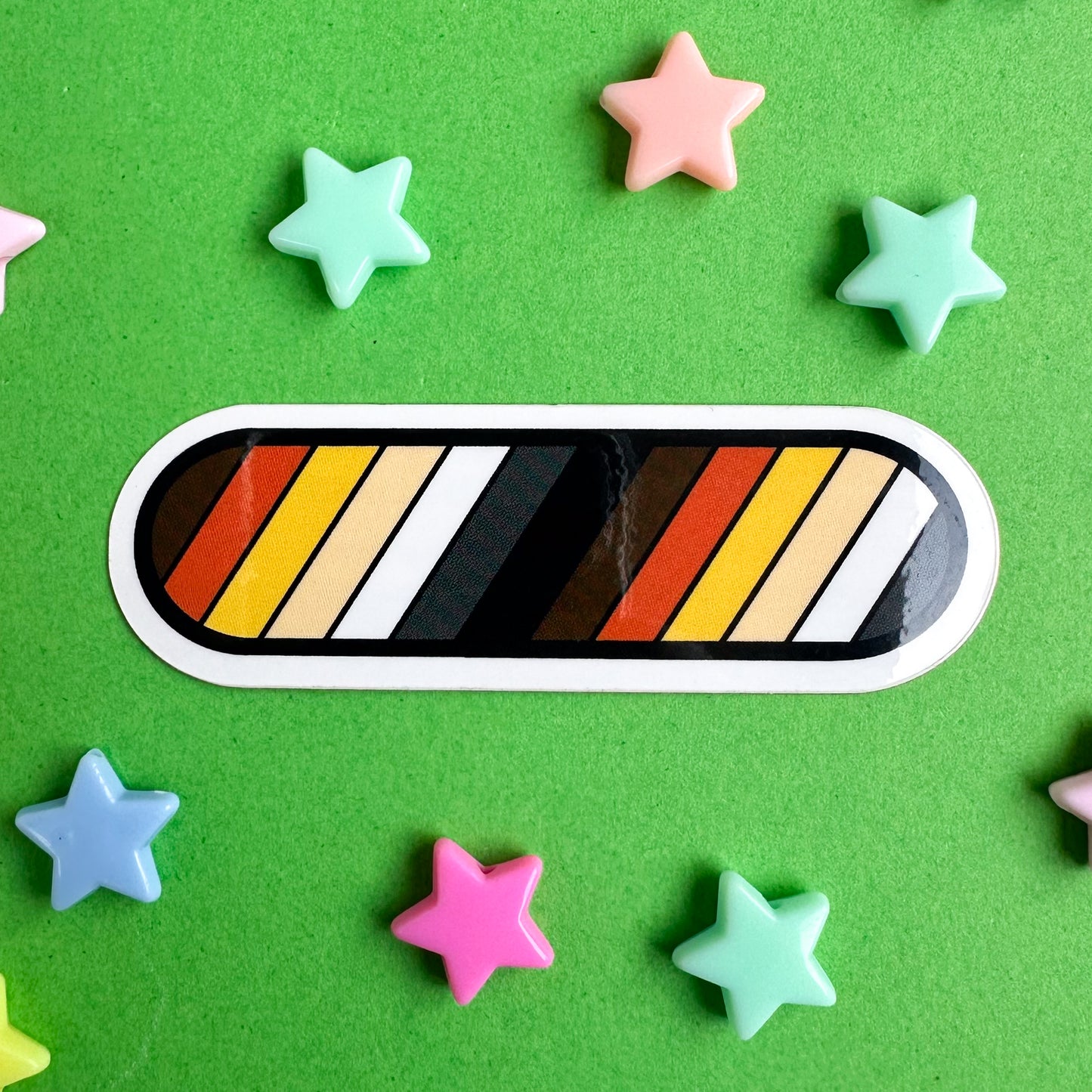A capsule shaped sticker with diagonal stripes in the colors of the Bear Pride Flag. The sticker is sitting on a green paper background with plastic star beads around it.