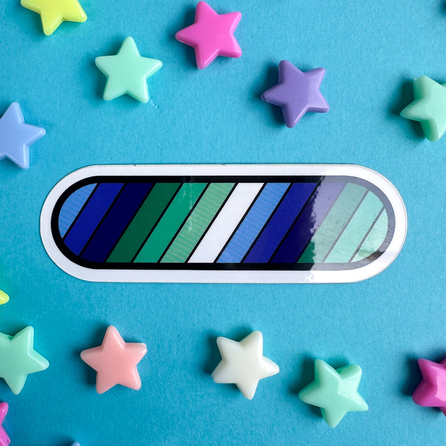 A vinyl sticker shaped like a capsule in the colors of the gay pride flag, blue and green. The sticker is on a blue background with star beads around it.