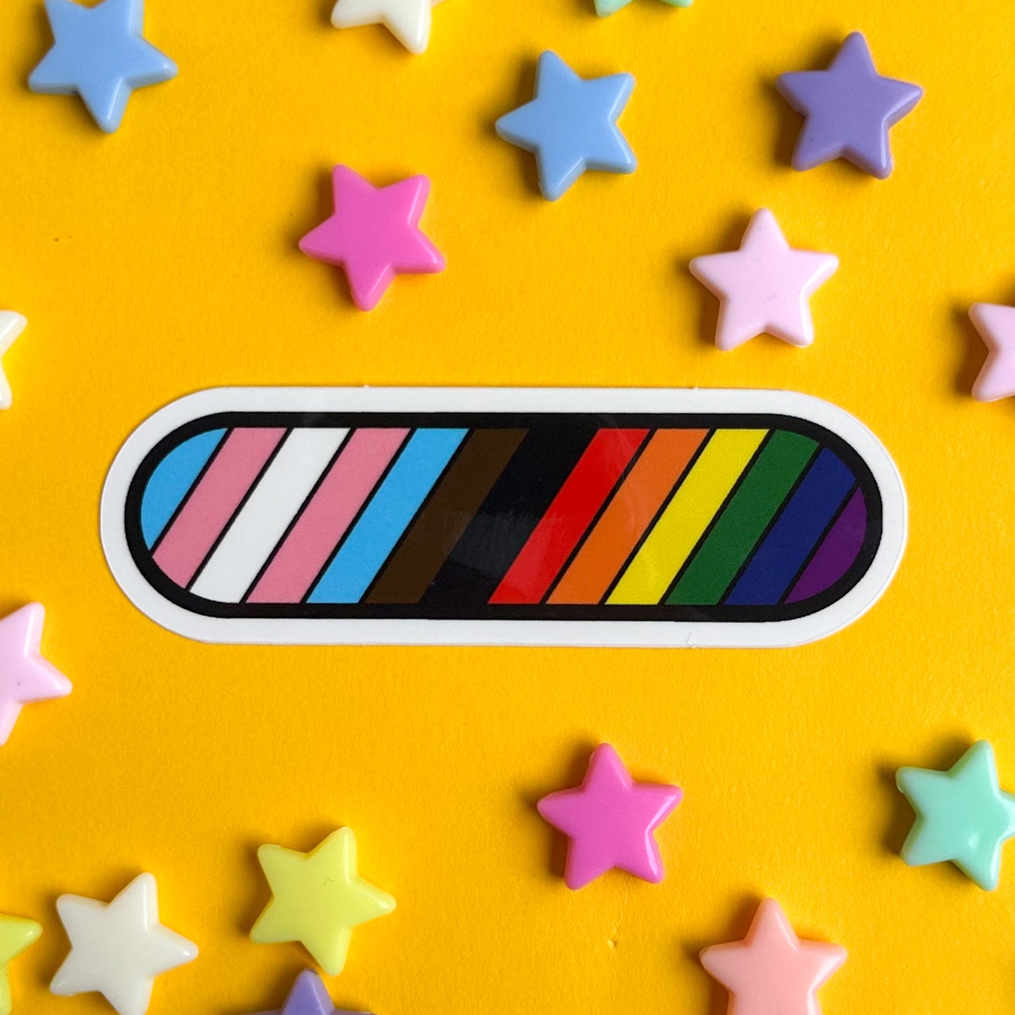 A sticker with diagonal stripes in the colors of the progress pride flag. The sticker is on a yellow background with pastel star beads around it.