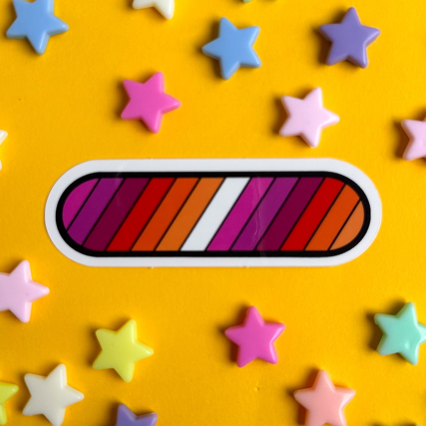An oval shaped vinyl sticker in the colors of the Lesbian Pride Flag. The sticker is on a yellow background with pastel star beads around it.