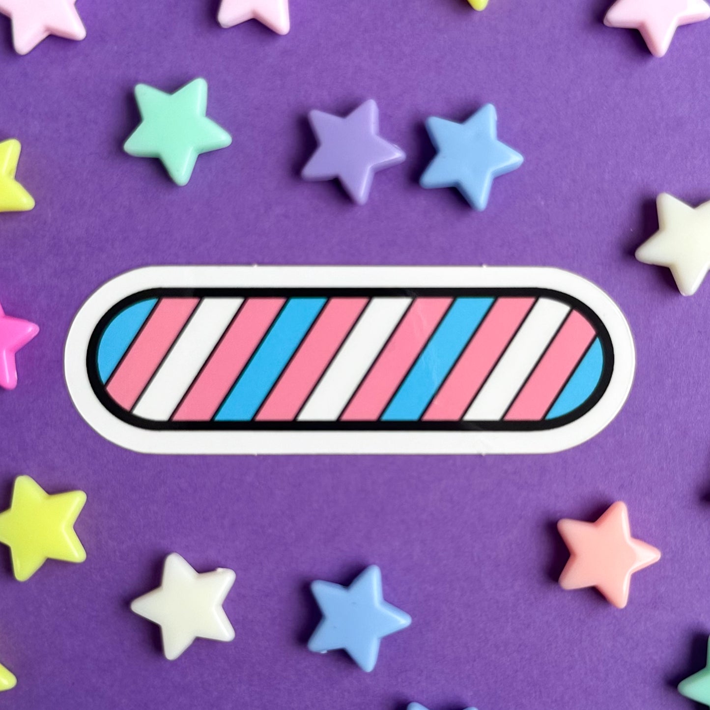 An oval shaped sticker with diagonal stripes of blue, pink, and white across it, the colors of the Trans Pride flag. The sticker is on a purple background with star beads around it.