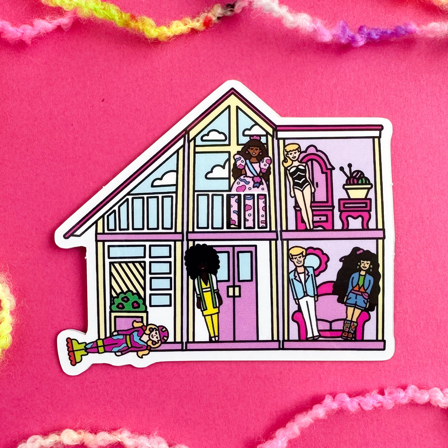A sticker shaped like a Barbie DreamHouse in pinks and creams. There are various Barbie toys strewn about the house including the original bathing suit barbie, ken, birthday barbie, rollerblade barbie and more. The sticker is on a pink background with fuzzy yarn around.