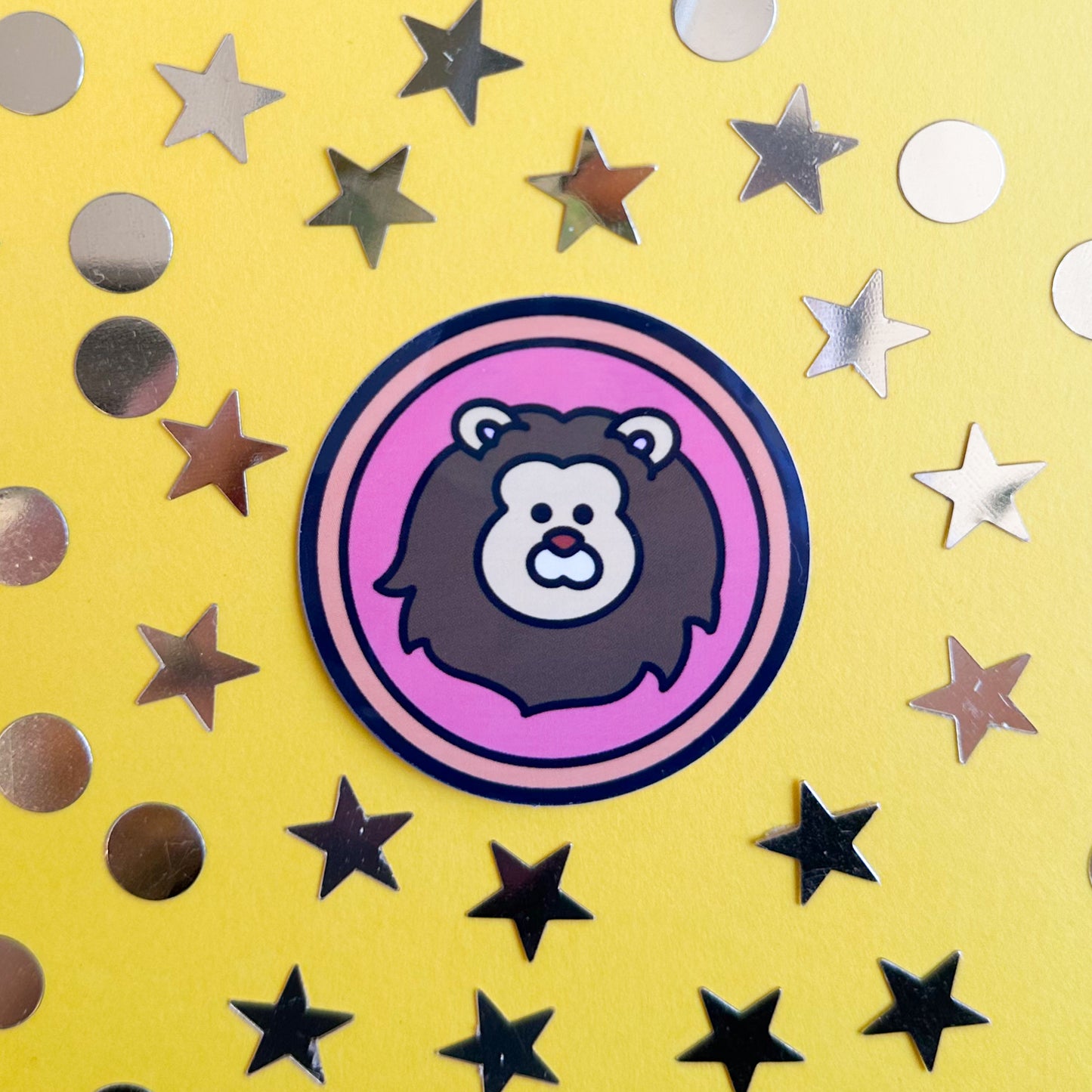 A circle sticker with a cute lion head on it to represent the Leo zodiac sign. The sticker is on a yellow background with gold star and circle confetti around it.