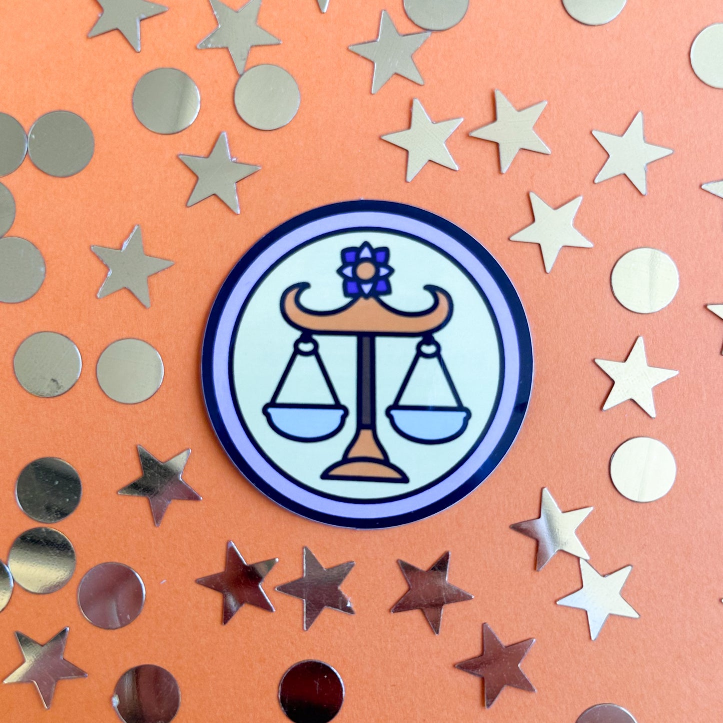 A sticker shaped like a circle with a set of scales in the circle to represent the Libra zodiac sign. The sticker is on an orange paper background that is covered in gold star and circle confetti.