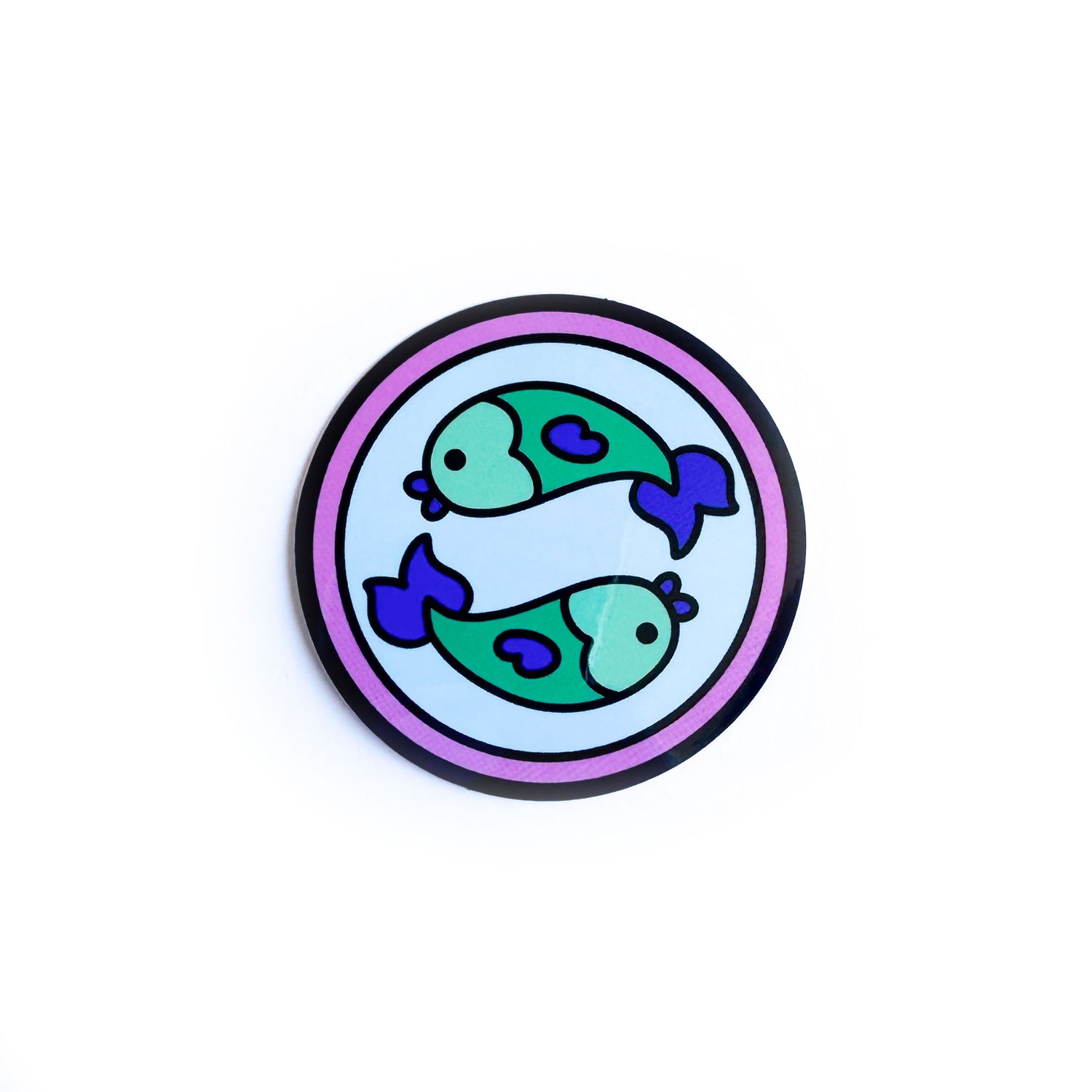 A sticker of a blue circle with a pink background with two green fish swimming in opposite directions on the blue circle to represent the Pisces zodiac sign.