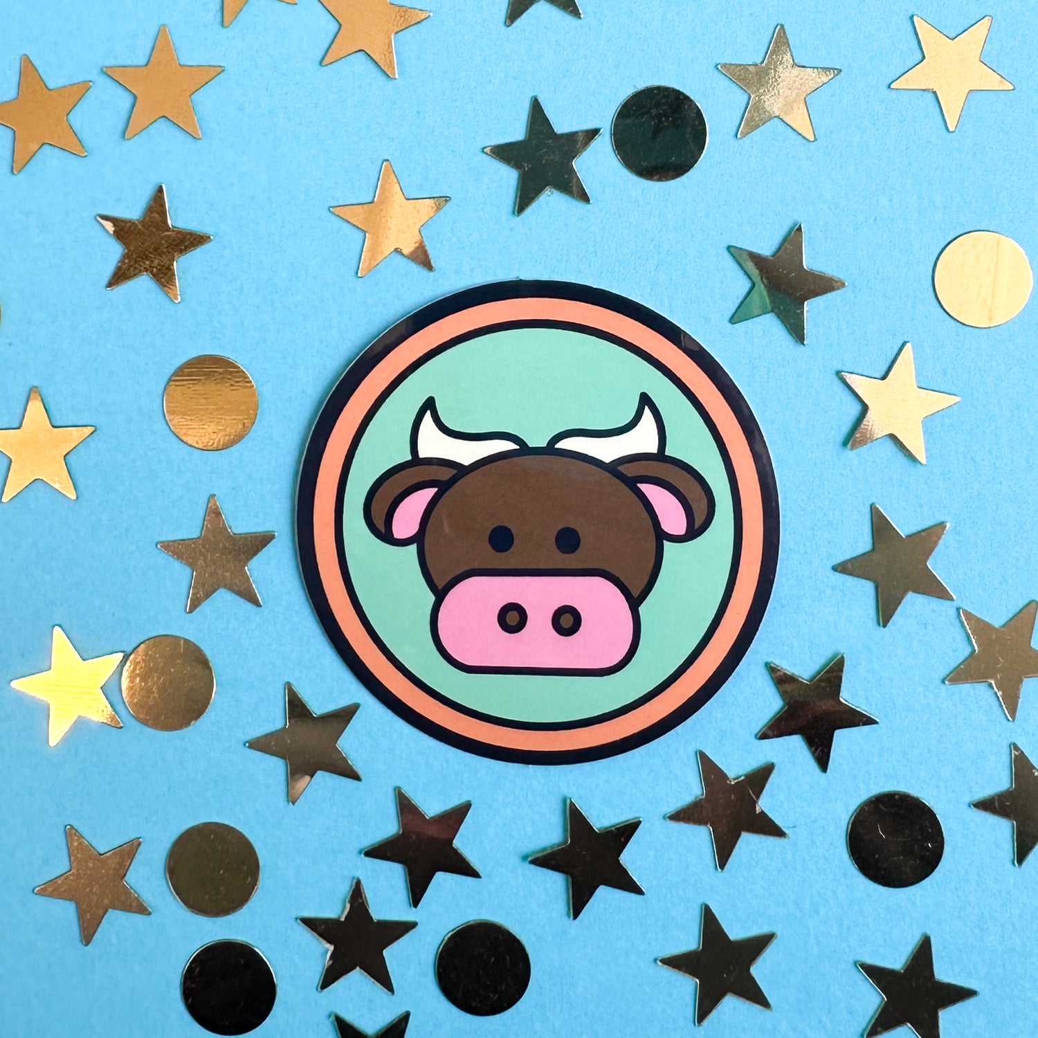 A circle sticker with a bull on it to represent the Taurus zodiac sign. The sticker is on a blue paper background that has gold confetti in star and circle shapes around it.