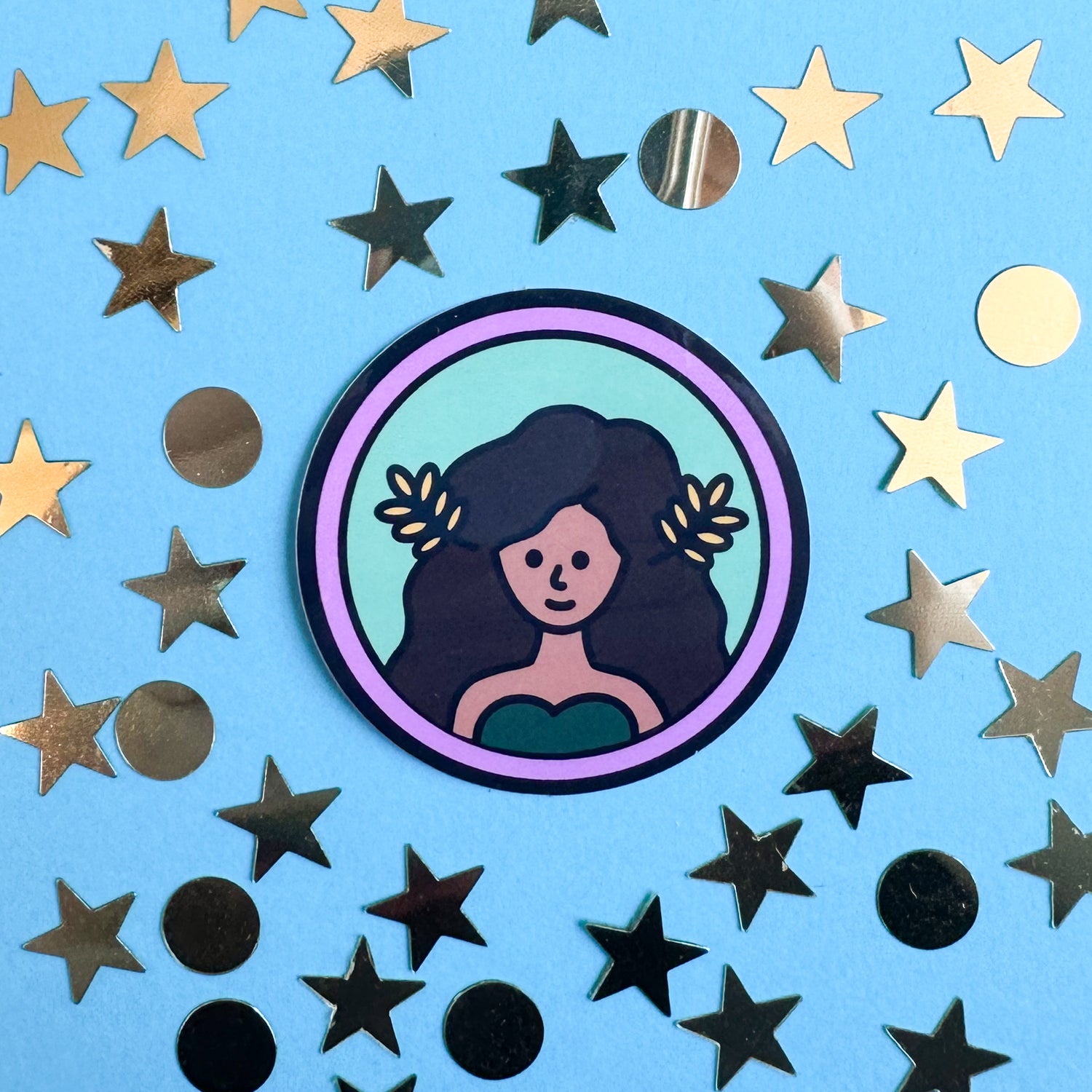 A circle sticker with an illustration of a woman with brown skin and flowing brown hair with wheat fronds in it. The sticker is on a blue paper background with gold confetti in circle and star shapes around the sticker.