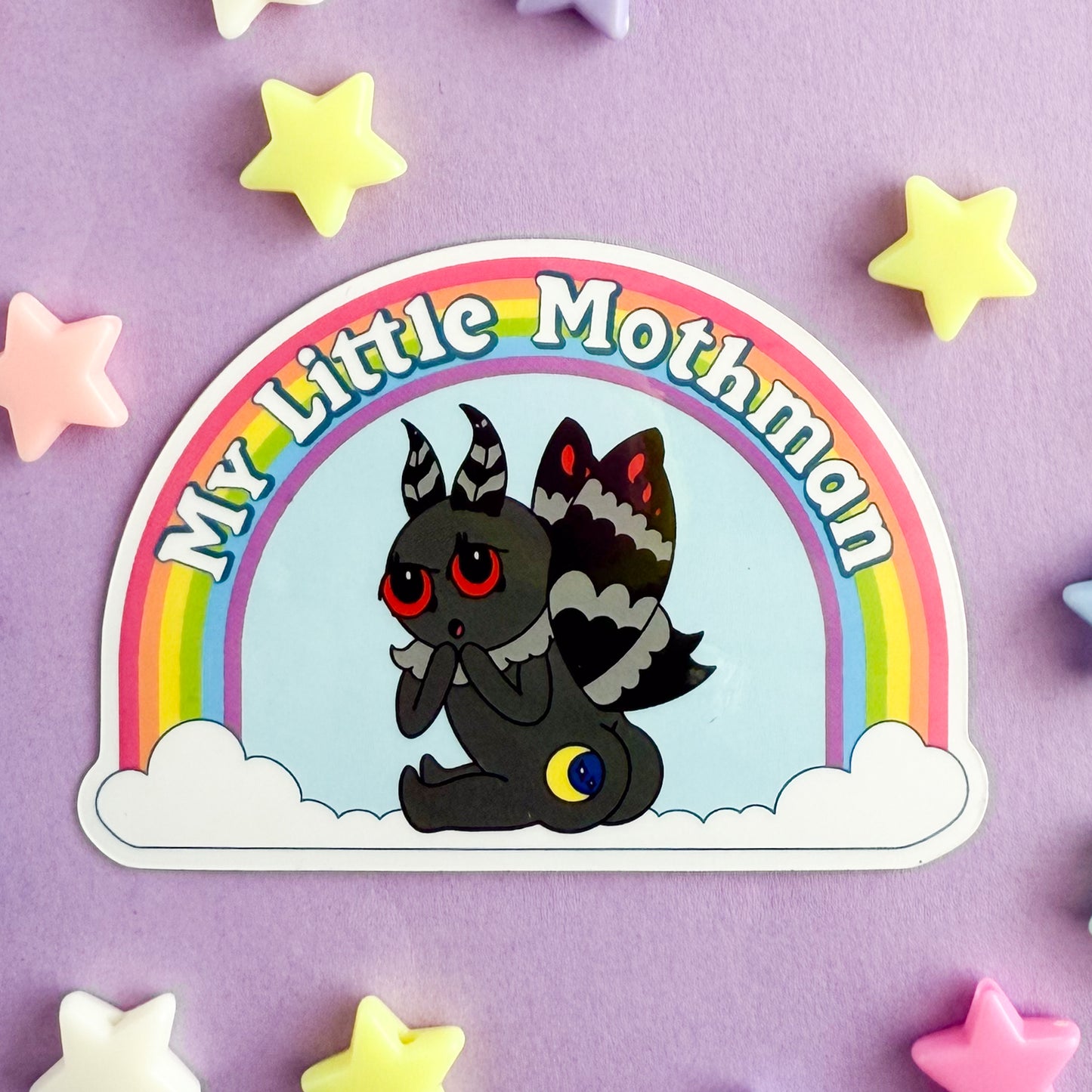 Sticker of a cartoon illustration of mothman turned with his butt towards you, his butt has a moon on one cheek. Above mothman is a rainbow with the words "My Little Mothman". The sticker is on a purple background with stars around it.