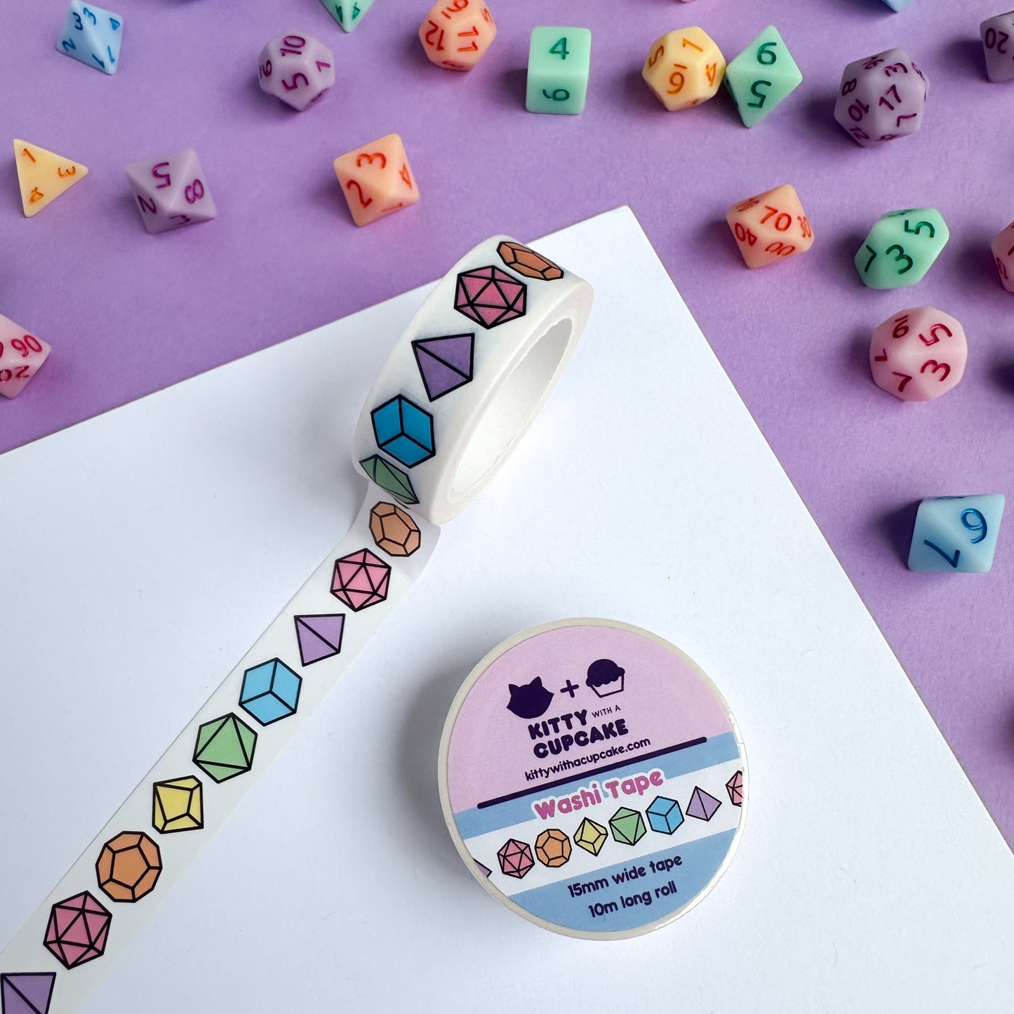 A roll of washi tape with illustrations of pastel rainbow dice on it. There are DnD dice in pastel colors that match the tape strewn around the piece of paper.