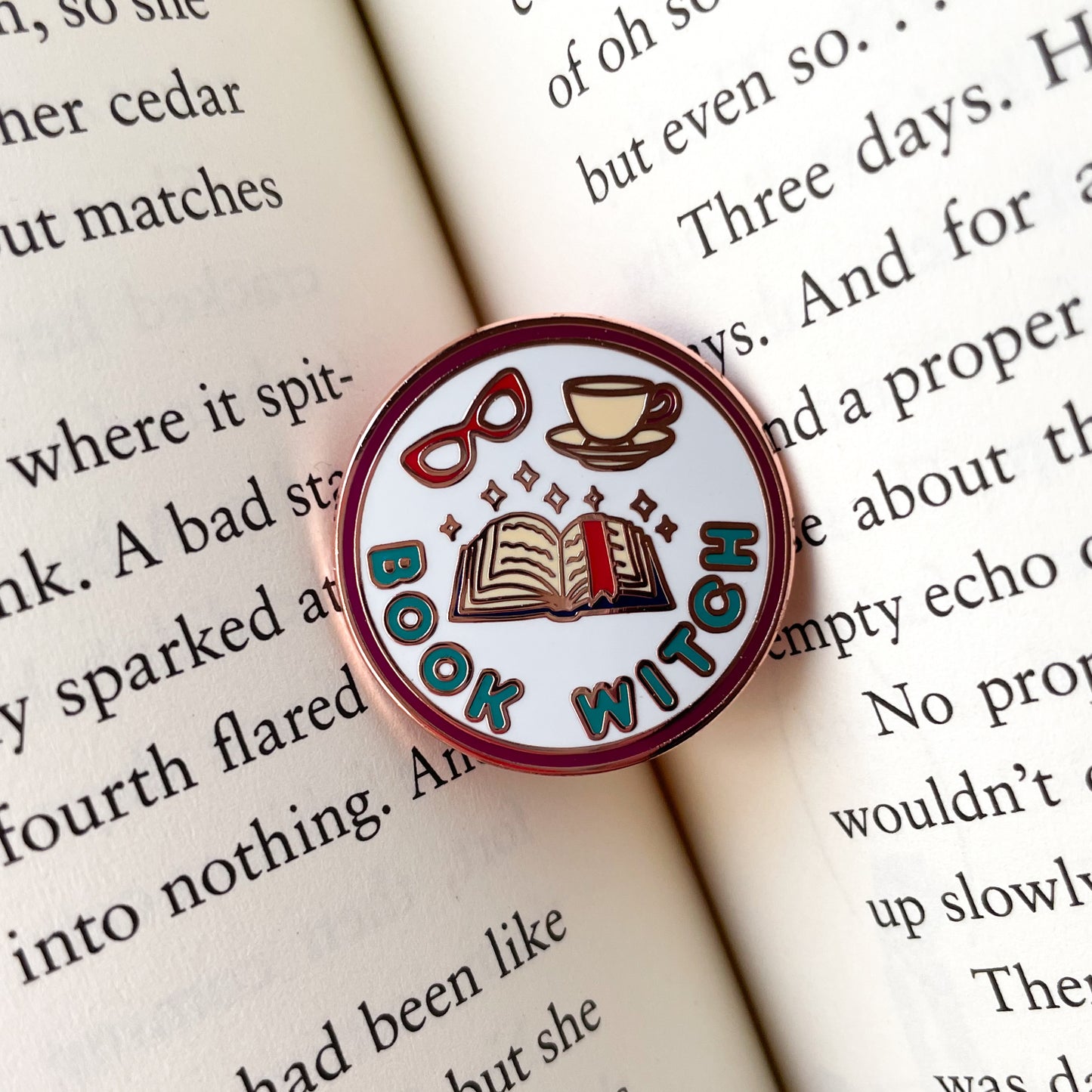 A circular enamel pin sitting in the middle of an open book. The pin says "Book Witch" and has an open book with a bookmark, sparkles, glasses and a teacup on it.
