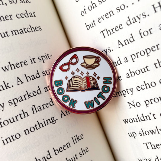 A circular enamel pin sitting in the middle of an open book. The pin says "Book Witch" and has an open book with a bookmark, sparkles, glasses and a teacup on it.
