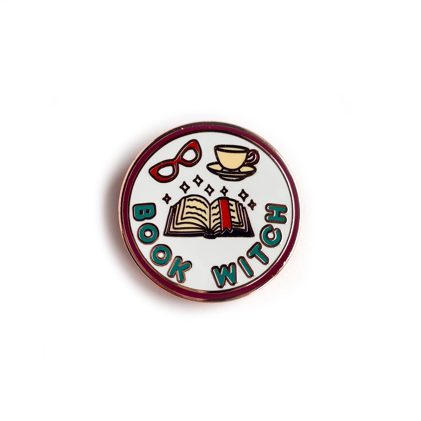 A circular enamel pin with copper metal details. It has a maroon border with a light blue background and turquoise words that say "Book Witch". Also depicted are an open book with a bookmark, a red pair of glasses and a teacup. There are sparkles above the book.