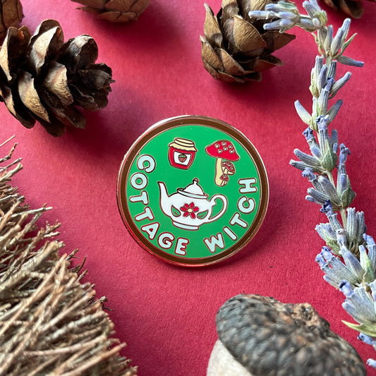 A circular enamel pin on a red background surrounded by small pine cones, an acorn, broom bristles, and a lavender sprig.