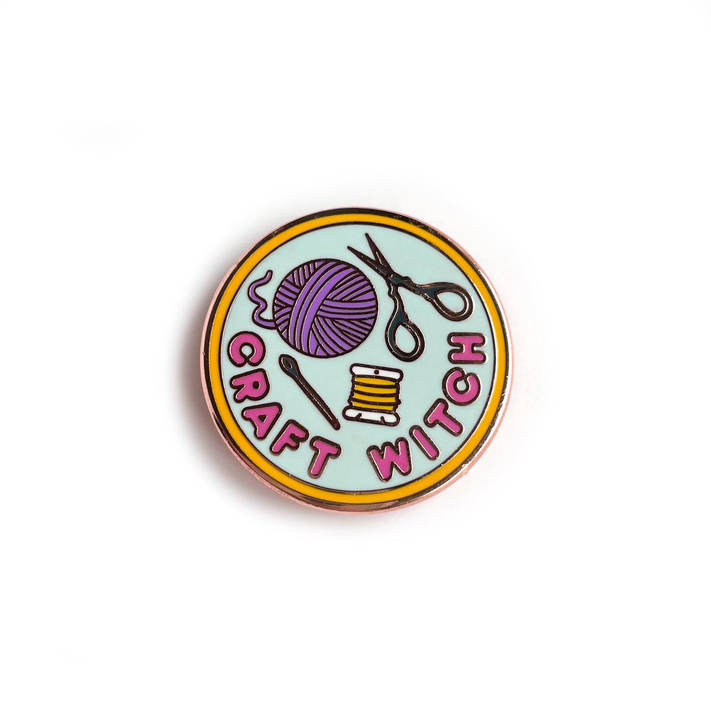 A circular enamel pin. It has a yellow border with a light blue background and hot pink letters that read "Craft Witch". It has a needle, embroidery floss, a purple yarn ball and scissors depicted on it.