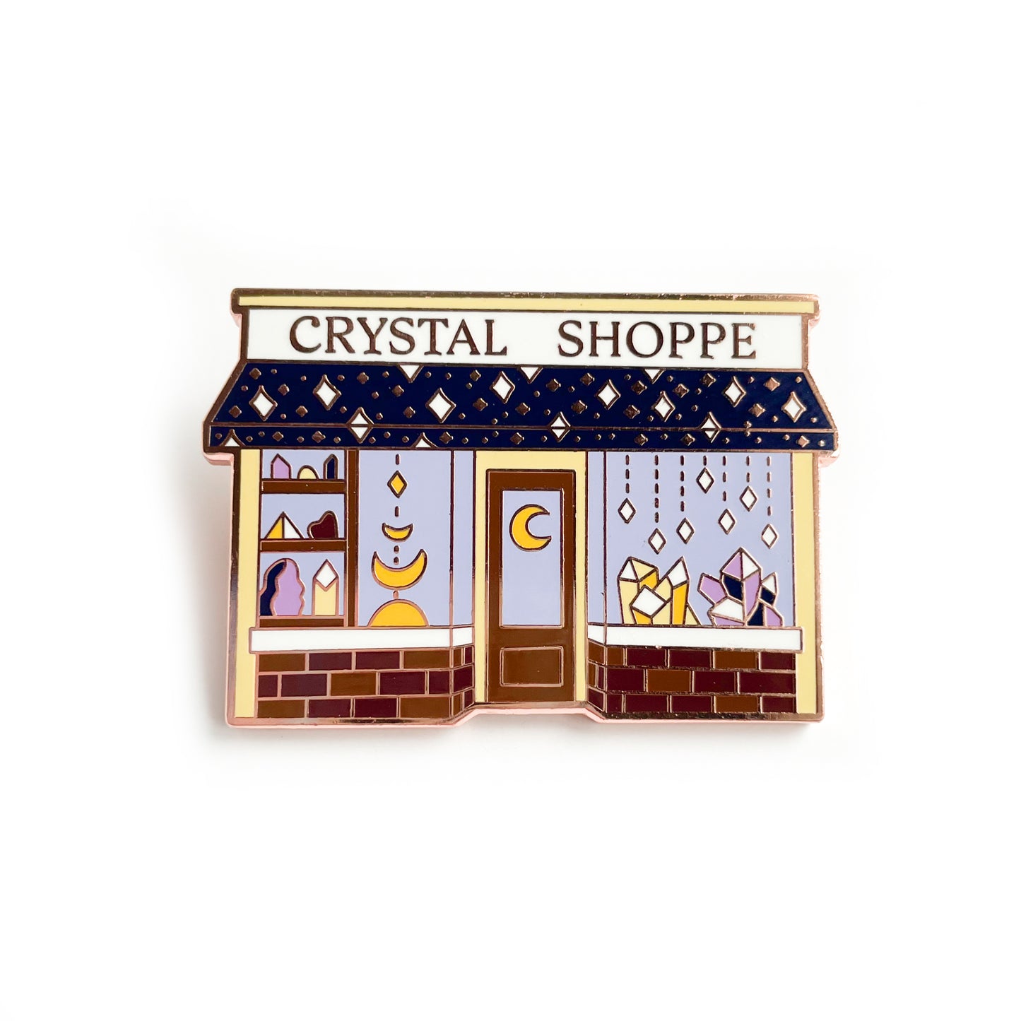 An enamel pin in the shape of shop with the words "Crystal Shoppe" on it. It has a door with a crescent moon on it. The windows are full of crystals in yellow and purple. The awning of the shop has sparkling stars on it and under the front windows are bricks.
