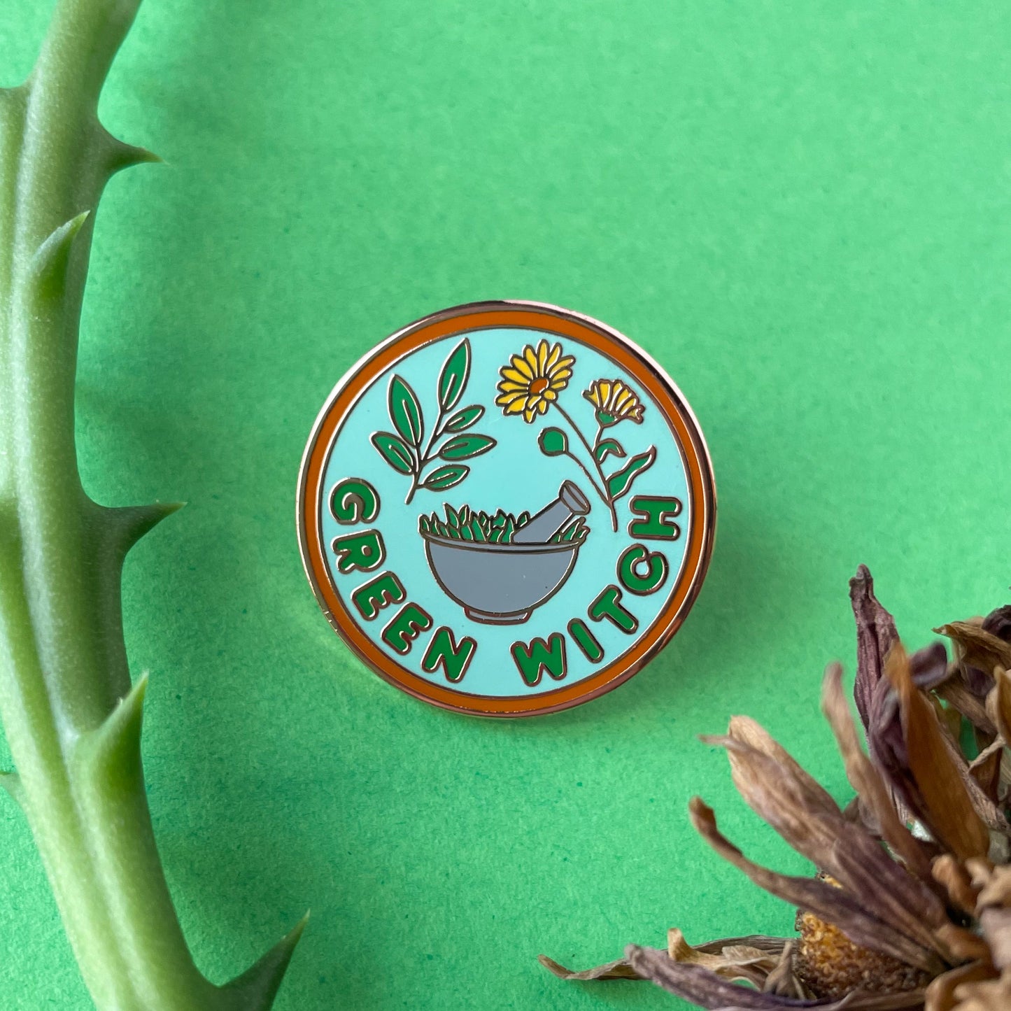 A circular enamel pin on a green background surrounded by dry flowers and a bit of succulent plant. The pin reads "Green Witch"