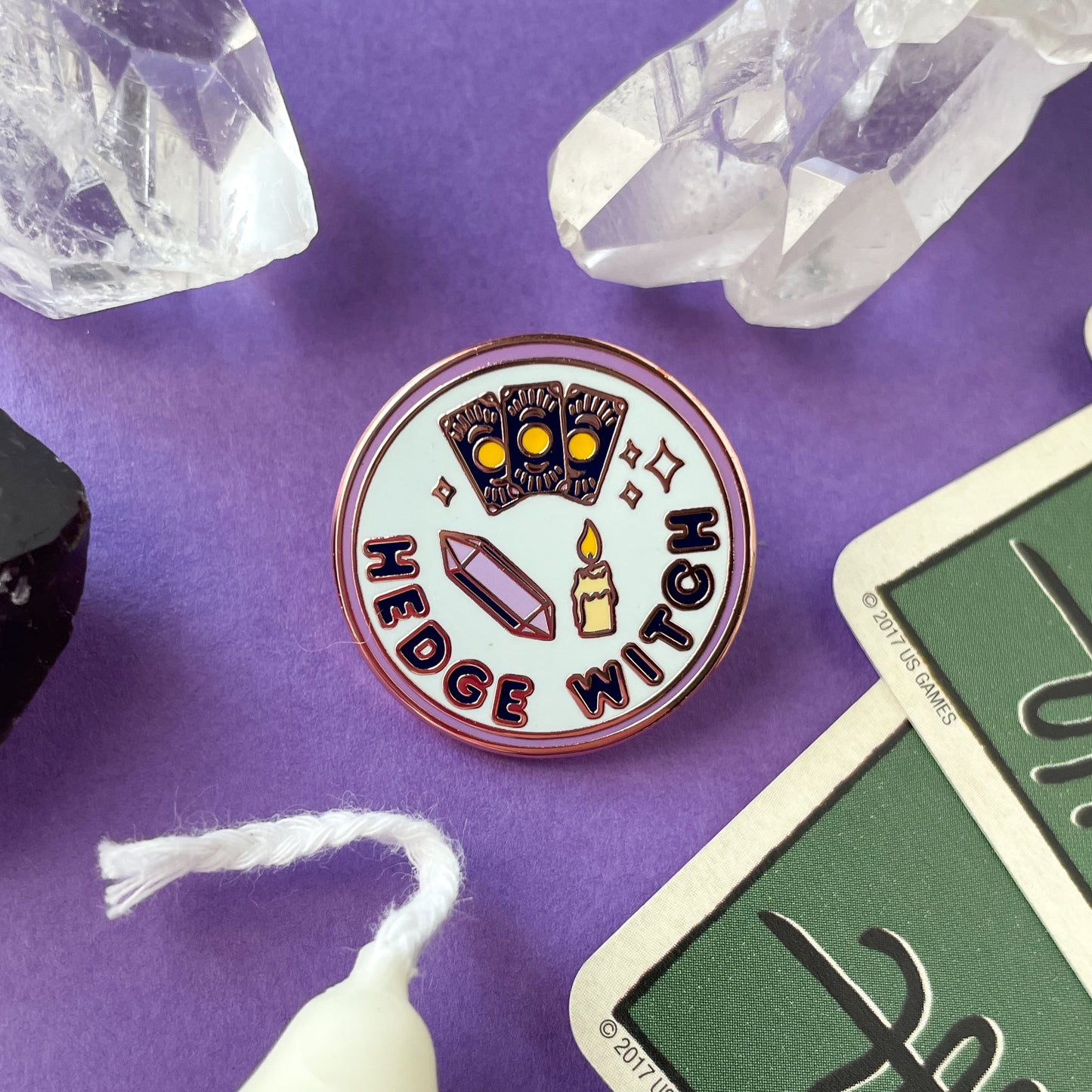 A circular enamel pin on a purple background surrounded by quartz crystals, cards and a candle. The pin reads "Hedge Witch".
