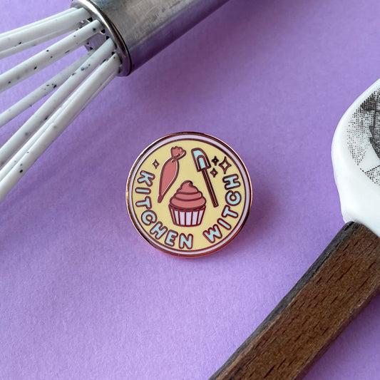 A circular enamel pin on a purple background surrounded by a whisk and spatula handle. The pin has the words "Kitchen Witch" on it.